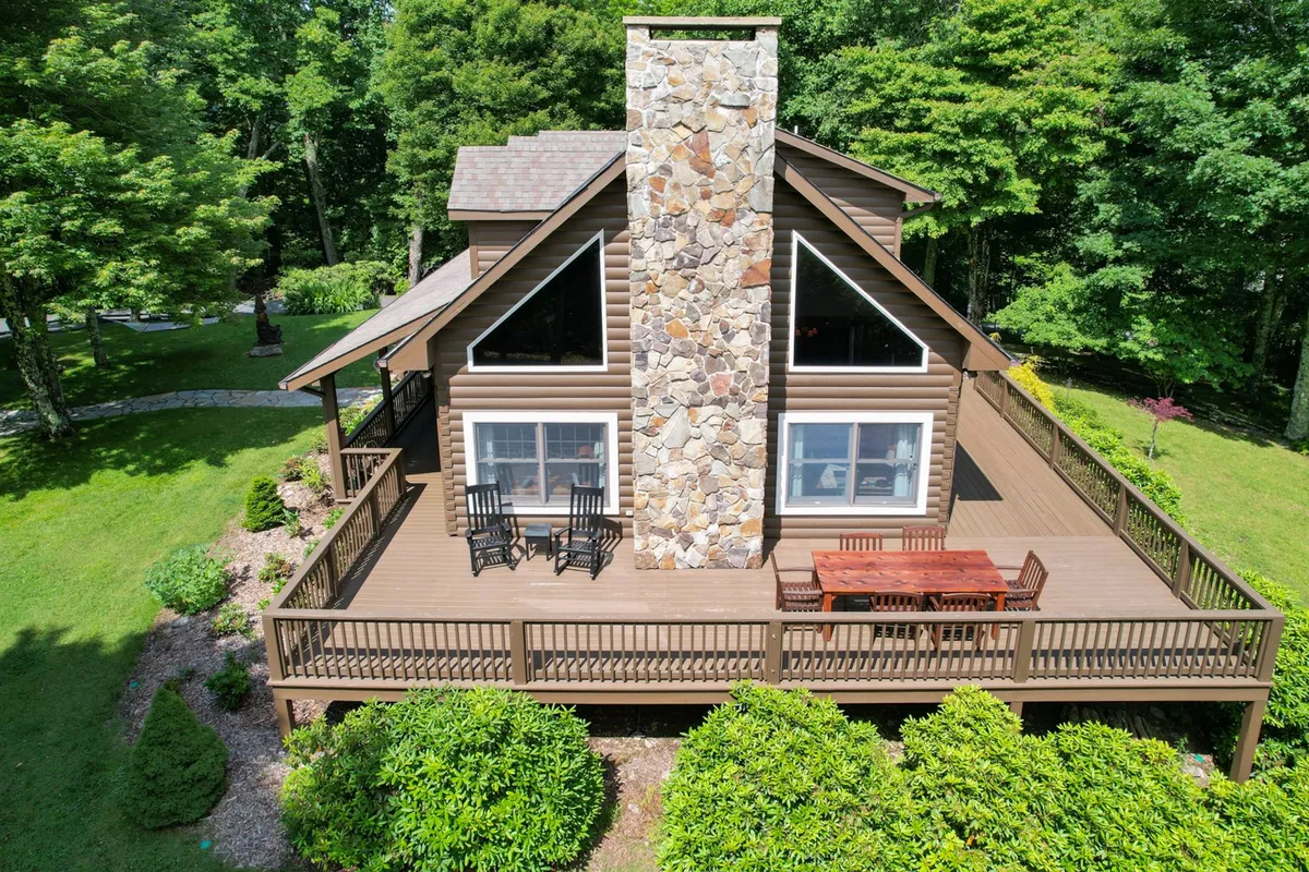 A large wooden deck with a dining area sits nestled in lush greenery, next to a stone chimney. The setting is inviting for outdoor dining, social gatherings, and spending tranquil sunset or evening hours with friends and loved ones.