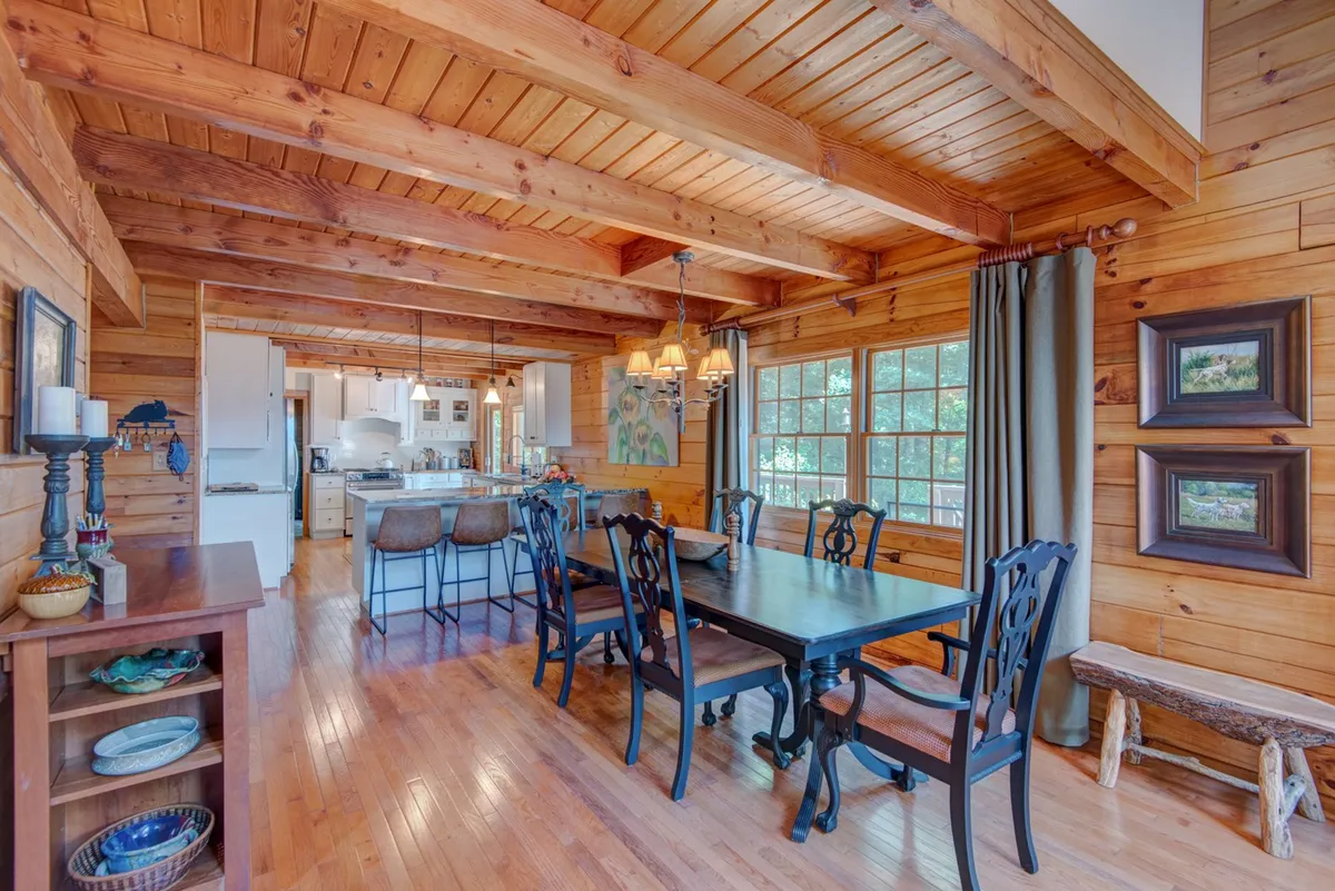 A spacious dining area with a wooden table set beneath a chandelier, adjacent to windows and with glimpses of the kitchen area. Wooden beams run overhead, and the setting feels intimate and inviting—ideal for family dinners, celebrations, or casual brunches.