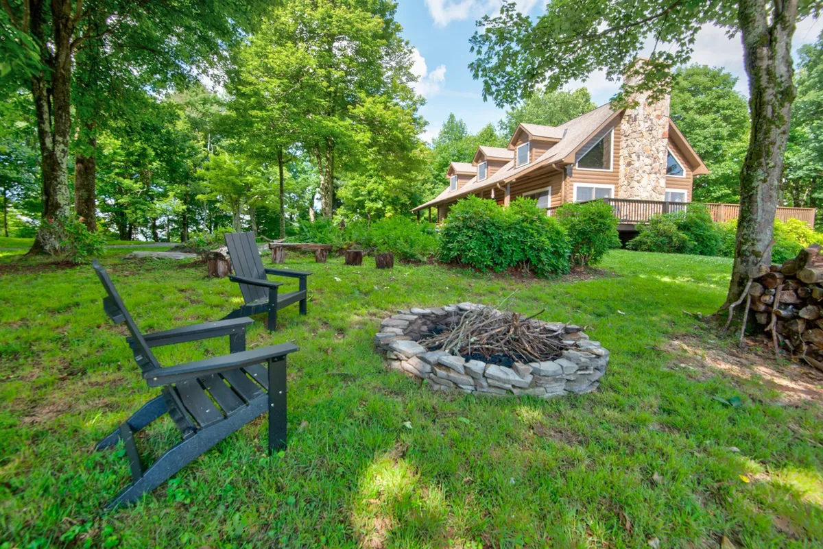 A cozy fire pit sits surrounded by classic Adirondack chairs, set on a lush green lawn with the stone house visible in the background. The area is enveloped by mature trees, creating a sense of privacy and natural seclusion. The scene suggests an inviting atmosphere for both daytime relaxation and evening gatherings around the fire under a starlit sky.