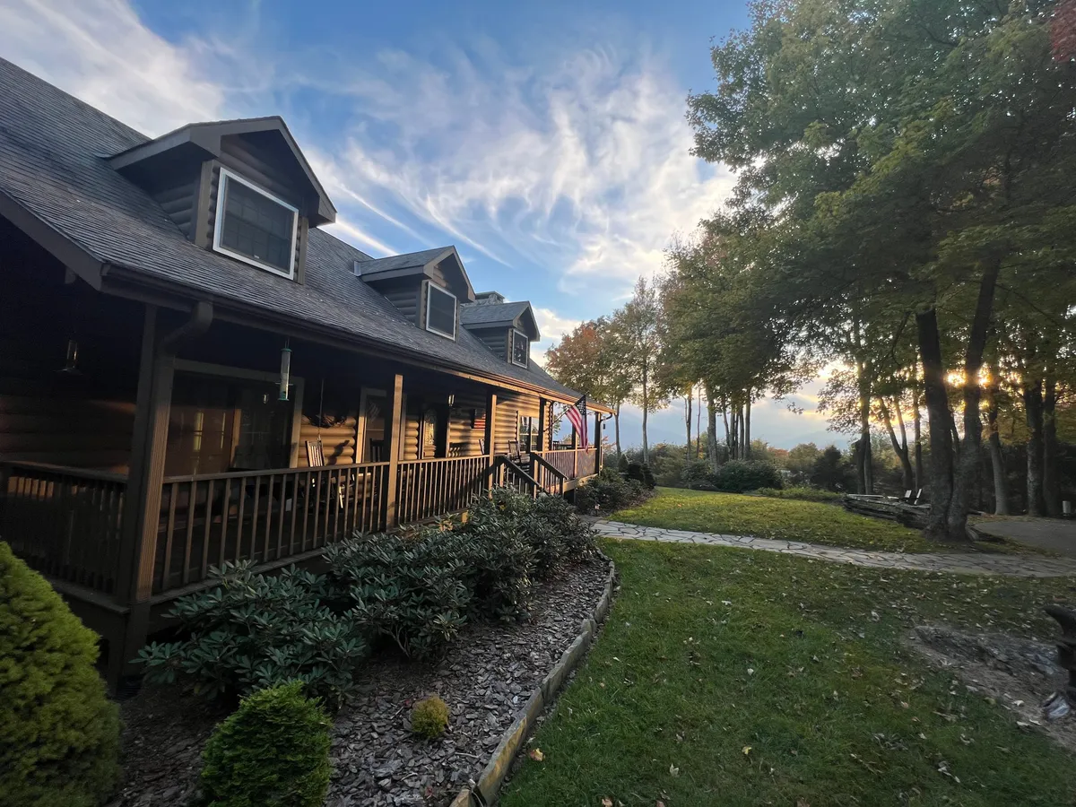 This image captures the property's log cabin structure with its expansive wrap-around porch, surrounded by lush greenery and bathed in the warm glow of sunset. The scene shows inviting outdoor spaces ideal for relaxing and absorbing the mountain atmosphere.
