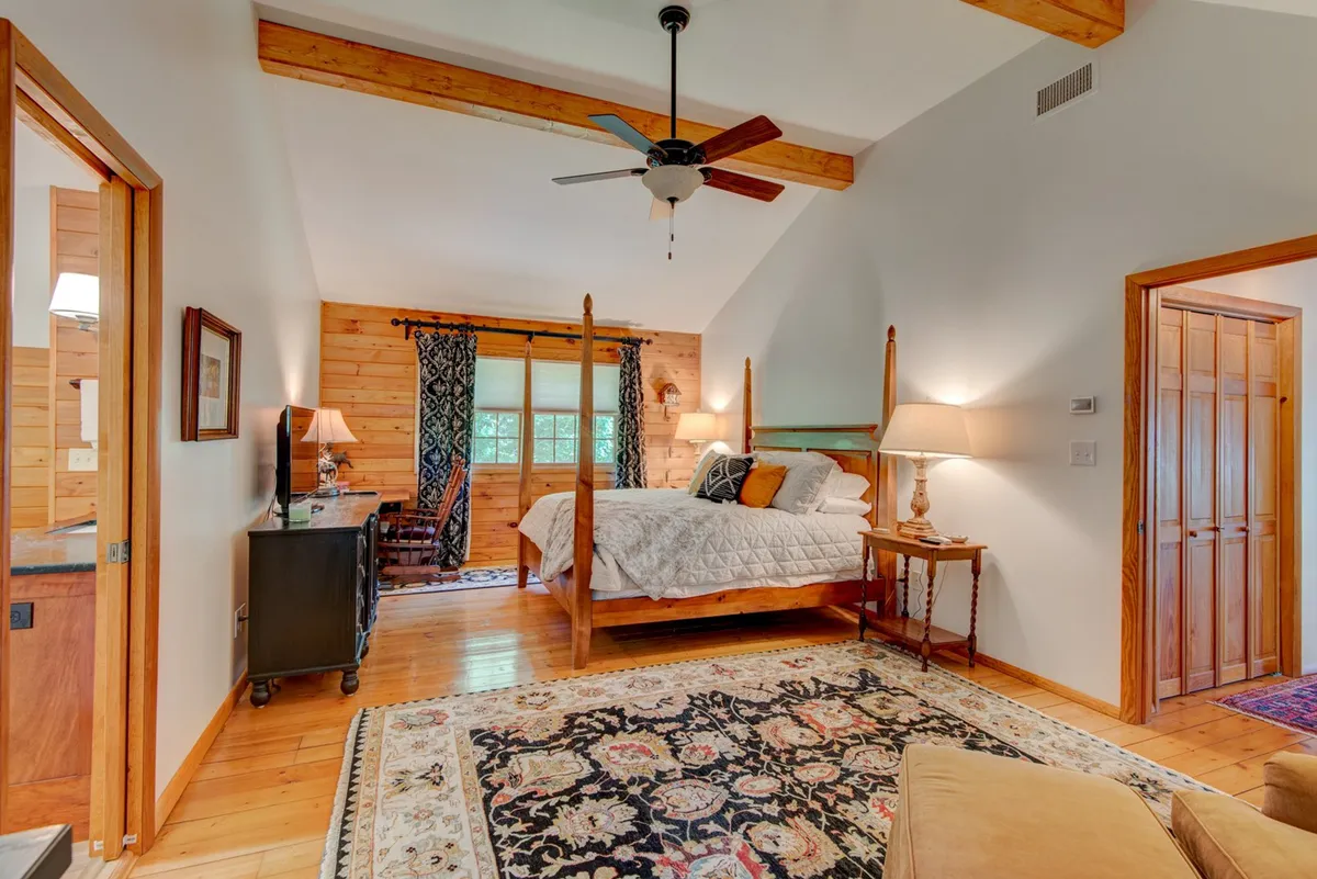 A cozy bedroom featuring a four-poster bed adorned with soft linens, a colorful area rug, wooden beams on the ceiling, and a window with curtains allowing natural light to softly illuminate the space. The room exudes warmth and relaxation, making it an ideal private haven.
