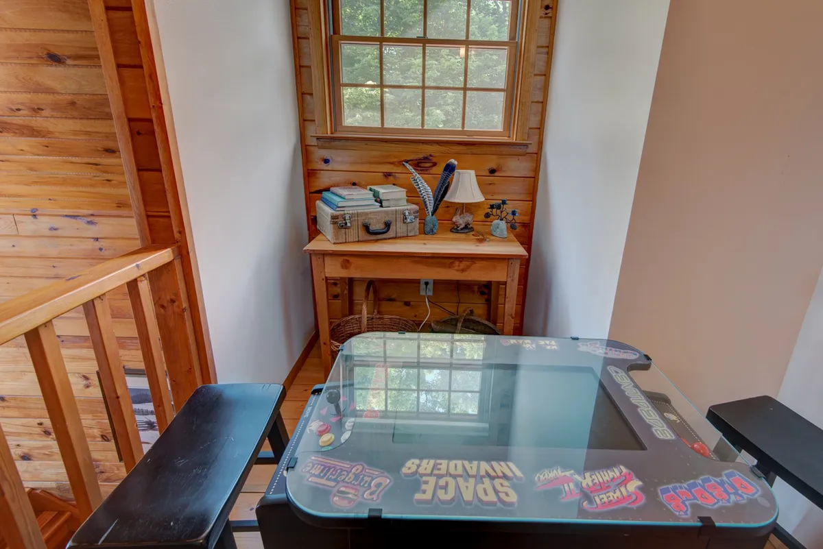 This image features a playful, sunlit loft area with a wooden desk, an arcade game table, and a window allowing in natural light. The inviting setup encourages both fun and relaxation, with ample space for seating and activities.