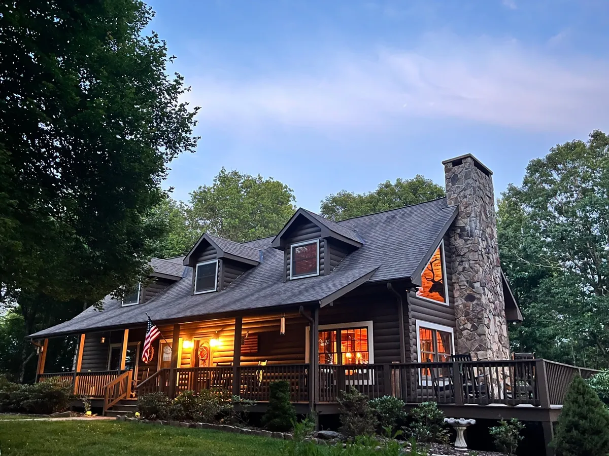 The image shows a charming log cabin perched on a private mountaintop lot, with a classic stone chimney, inviting front porch lined with a wooden railing, and windows glowing warmly from within. An American flag ripples gently in the mountain breeze, enhancing the sense of welcome. The porch offers panoramic mountain views and ample space for relaxing or gathering with loved ones.