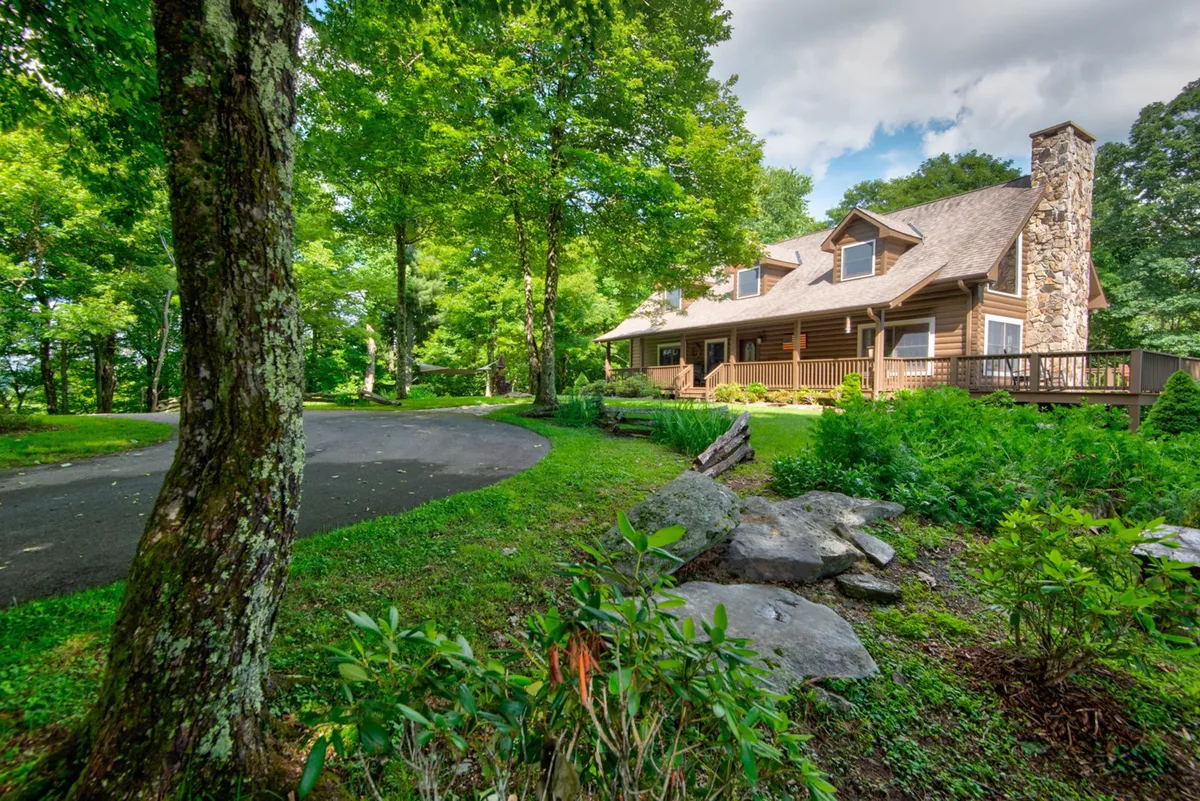 The charming log cabin is nestled among lush greenery, with a curved driveway and a prominent stone chimney. The image radiates peacefulness and highlights the privacy and seclusion offered by the property.