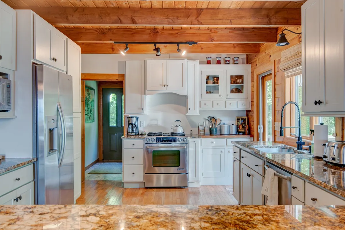 A beautifully remodeled kitchen with gleaming granite countertops, stainless steel appliances, wooden beams overhead, and large windows letting in abundant natural light. The kitchen is open to the rest of the living space, highlighting the flow between cooking and socializing.