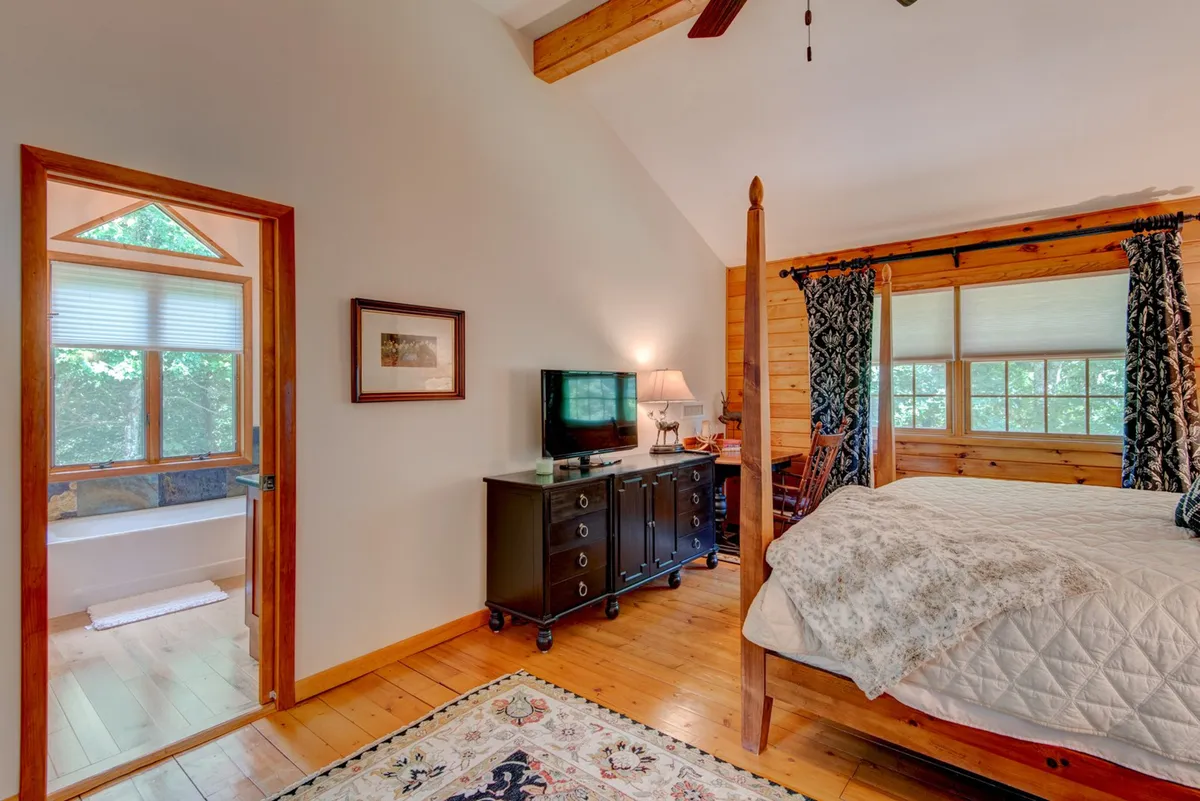 A tranquil bedroom with a four-poster bed, chest of drawers, a large window letting in views of nature, and a decorative rug. The overall atmosphere is calm and restful, suggesting a space designed for relaxation and rejuvenation.
