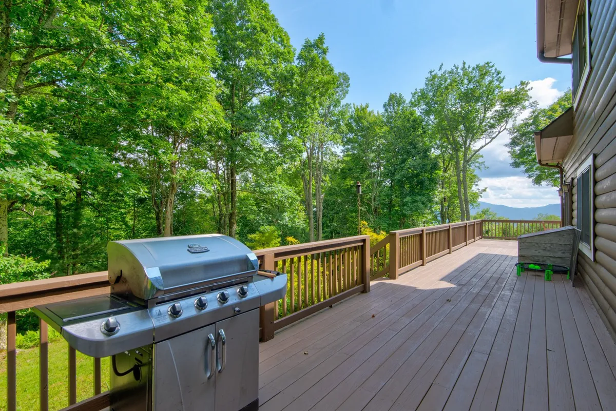 An expansive wrap-around deck with a grill overlooks a tranquil wooded setting. The deck is bordered by sturdy wooden railings and surrounded by trees, offering both privacy and panoramic forest views. The grill hints at outdoor cooking and entertaining, while the spacious deck invites relaxation in the fresh mountain air.