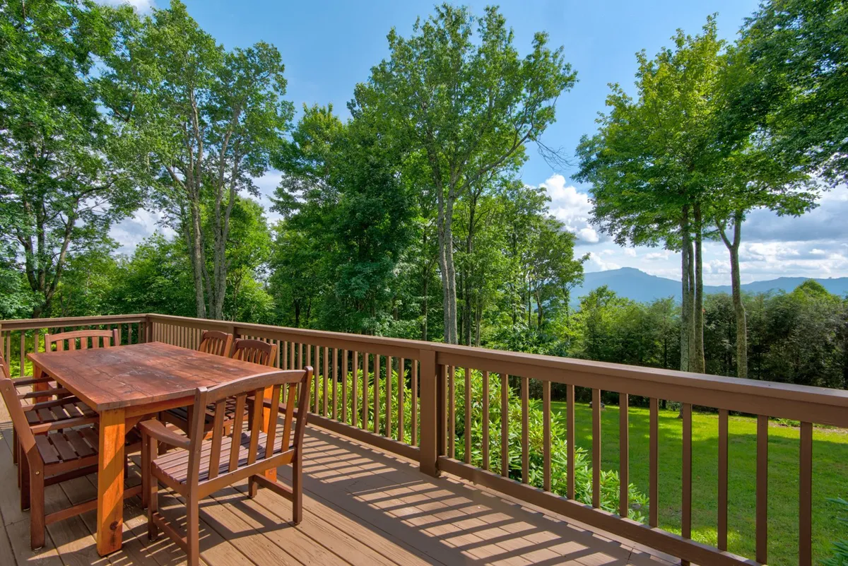 A spacious wooden dining table sits on an outdoor deck surrounded by lush trees, with a clear view of distant mountains. The setup invites outdoor dining and relaxation, all set against a tranquil mountain backdrop.